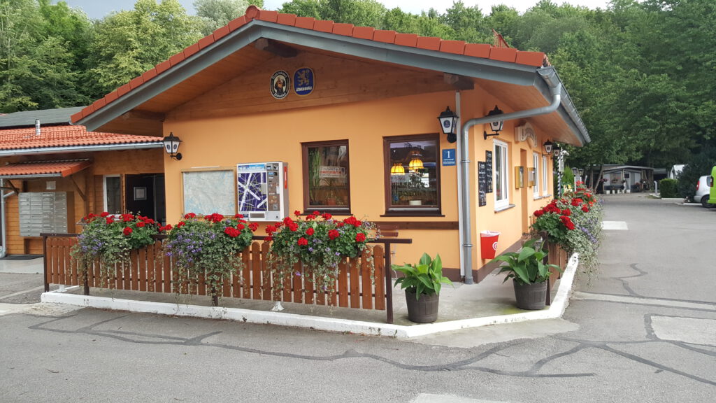 Vorderansicht der Campingstubn, von vielen Pflanzen umgeben. Auf zwei Seiten des einstöckigen Hauses befindet sich eine asphaltierte Straße im Campingbereich.