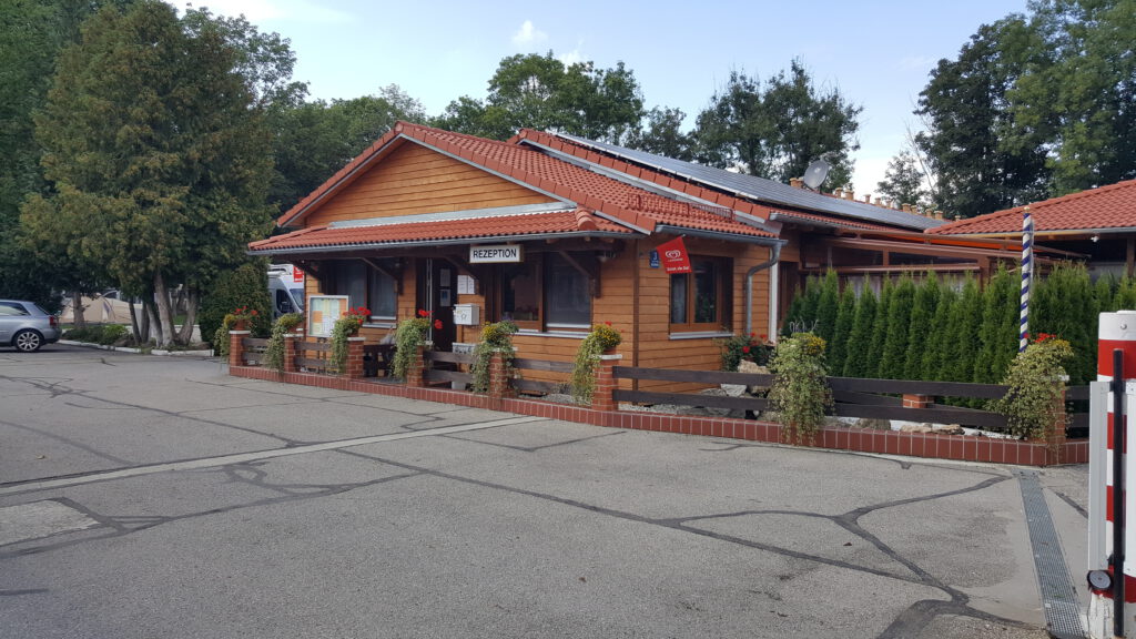 Das Rezeptionshaus, eingezäunt und von vielen Pflanzen umgeben. Davor eine asphaltierte Fäche, im Hintergrund sind Bäume zu sehen.