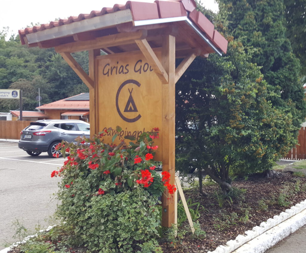 Ein überdachtes Willkommensschild, etwas von roten Blumen überwachsen, auf einem Grünstreifen beim Parkplatz. Die Aufschrift lautet "Grias God - Campingplatz Nord-West".