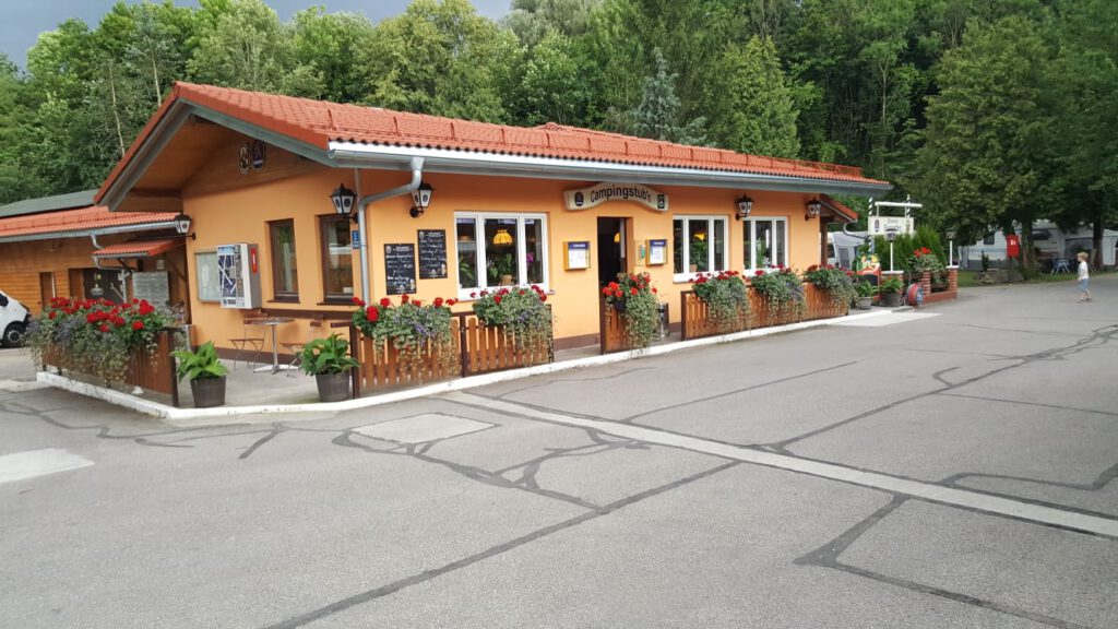 Campingstubn Frontansicht mit Eingang. Das einstöckige beige-farbene Haus hat viele Fenster. An der Wand sind Laternen montiert. Es ist umgeben von einem reichhaltig begrünten Holzzaun mit mehreren Durchgängen.