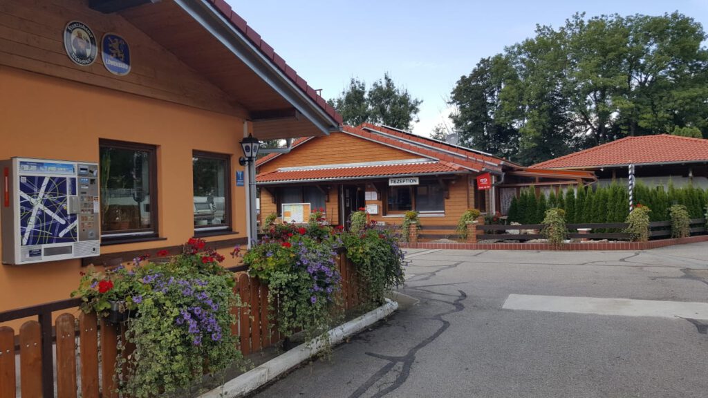 Campingstubn Seite mit Zigarettenautomat links im Bild, gegenüber das Rezeptionsgebäude. Rund um die Campingstubn ein Holzzaun mit überbordenden Blumenkästen mit viel Grün und roten und violetten Blüten.