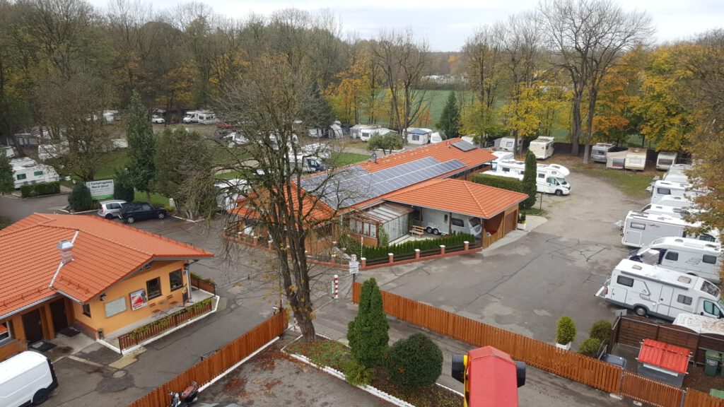 Luftaufnahme vom Campingplatz. Links ist die Campingstubn zu sehen, zentral ist ein weiteres Gebäude mit orange-farbenem Dach und großer Photovoltaik-Anlage. Rundherum sind die Stellplätze für Wohnmobile und Wohnwägen. Zwischen den Gebäuden und Stellplätzen befinden sich asphaltierte Straßen sowie zahlreiche Bäume.